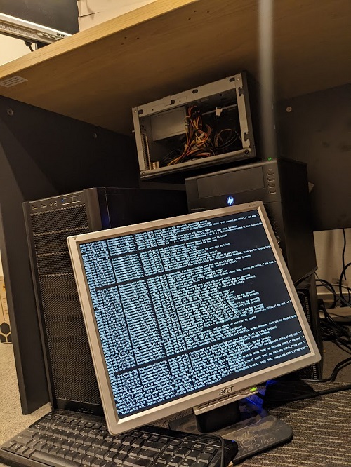 An old 4:3 ratio LCD computer screen sits yunder a desk in front of a few old headless computers, some partially deconstructed. A terminal takes up the entire screen, displaying linhe after line of logging information.