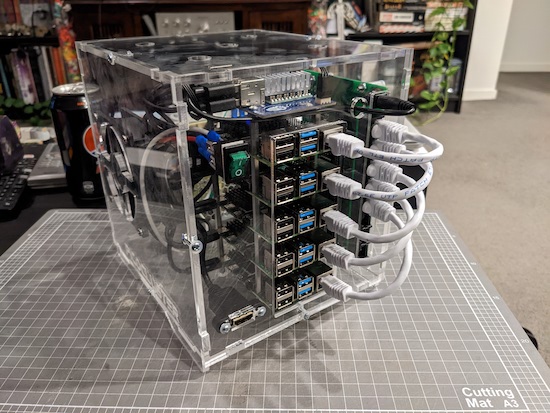 An assembled PicoCluster Raspberry Pi cluster computer. It consists of transparent acrylic panels housing a fan on one side, a network switch on the other, and five Raspberry Pi computer stacked on top of each other with spacers. A solid looking green rocker power switch on the front sits above a single HDMI port.