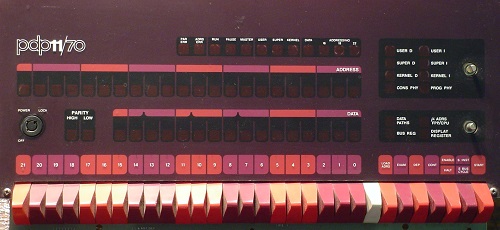 The front panel of a PDP-11/70 computer with rows of blinkenlights, a key to switch it on or off, and a row of about 30 switches to handle the output and debugging of the machine. It’s in a beautiful mauve, purple, and orange colour scheme.