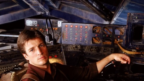 A handsome Malcolm Reynolds played by Nathan Fillion sitting at the control seat, behind him a dizzying array of switches and buttons and controls to pilot his spacecraft.