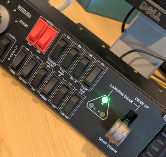 A flight simulator panel with various toggles and dials. In a section marked Landing Gear the top of a triangle of three lights is glowing bright green with a visible lens flare.