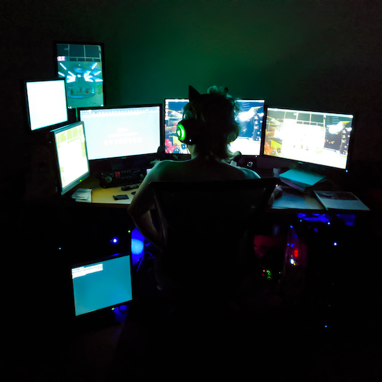 A photo of Az from behind, lit entirely by the glow of a collection of seven monitors. Six are strewn across their desk, one sits below it, all showing various desktops and terminals.