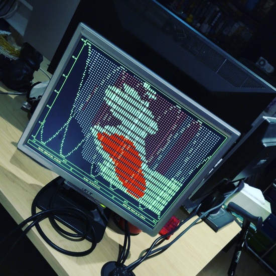 An old 4:3 ratio Dell LCD computer monitor sits on a desk, showing a terminal interface using limited character and colour palettes to render rainfall mapped over the local area. Bright green squares represnt some rain in the last five minutes, bright red squares representing heavy rain in the last five minutes. Duller green and red dots represent light and heavy rain detected in the 15 minute period prior. Two sine waves oscillate across the screen for no discernable purpose and some labels down the bottom give information about the time, temperature, and wind.